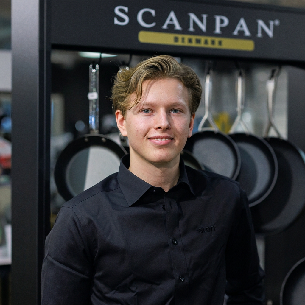 Young man in a black button-down shirt stands in a cookware showroom with hanging pots and a ScANPAN sign above.