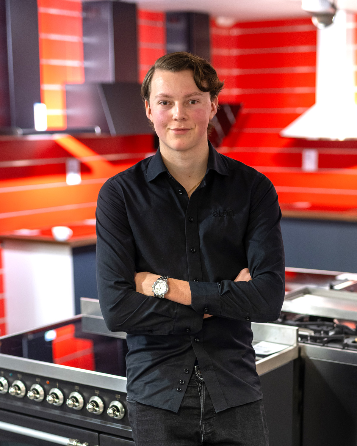 Portrait of a young man with arms crossed, wearing a dark shirt, standing in a red, modern kitchen showroom with appliances behind him.